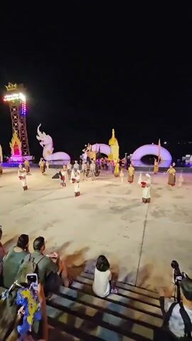 This video captures a nighttime cultural performance in Ko Kaeo, Thailand. The scene is set on a large, flat, paved area, likely a plaza or outdoor stage, under a dark night sky. In the background, a prominent, brightly lit tower with colorful lights stands tall. A large, white, serpent-like sculpture, possibly representing a Naga, is a central element of the stage design, curving across the performance area and incorporating decorative golden structures.

On the performance stage, several individuals dressed in traditional costumes are engaged in what appears to be a dance or ceremonial procession. Their outfits are predominantly white with red accents around the bottom of their garments and headwear. Some performers hold long, slender objects. A horse-drawn carriage is also visible on the stage, adding to the sense of historical or ceremonial context. The overall atmosphere is festive and visually striking, with the lighting from the tower and stage decorations creating a vibrant spectacle against the darkness.

In the foreground, a group of spectators is seated on tiered steps, watching the performance. Most are facing the stage, with some individuals holding up their mobile phones, presumably to record the event. The presence of cameras and the engaged audience suggests this is a public event or festival. The weather appears clear, as there are no visible signs of rain or harsh conditions. The mood seems to be one of enjoyment and appreciation for the cultural display. No legible text is visible in the video.