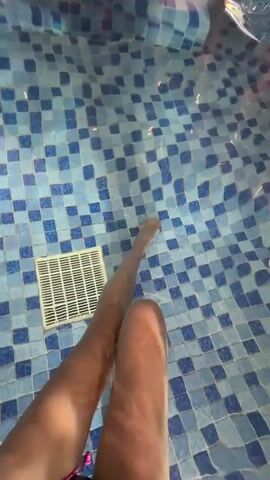 In an indoor swimming pool, a person's legs are submerged in the foreground, extending towards a white drain cover on the blue and white tiled bottom. Another person's legs and lower torso are visible in the background, floating or swimming on the water's surface. The water is clear, reflecting light. The scene depicts a relaxed moment in the pool.