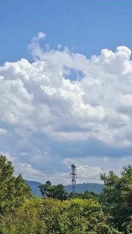 This is a vertical video of a sky with clouds. The clouds are white and fluffy, and they are against a blue sky. There are some trees and a transmission tower in the foreground. The video is mostly static, but there are some slight movements of the clouds and the camera.