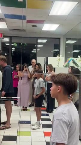 A group of people are inside a brightly lit building, possibly a store or event space. A mirror reflects the group, allowing for a doubled view. The group includes men, women, and children of varying ages. Some are taking pictures or holding phones. Most are standing and observing something happening. The environment includes a patterned floor with colorful squares, walls with reflective surfaces, and some decor with drawings. The focus is on a young boy in the foreground wearing a grey shirt, black shorts, and white Crocs.
