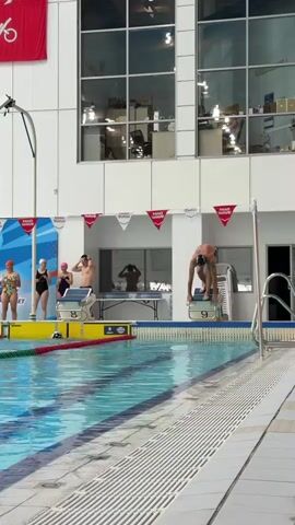 A swimmer dives from a starting block into a swimming pool. The swimmer is in mid-air, performing a flip. There are several other swimmers and officials visible on the pool deck. The pool is indoors, with a large window in the background showing offices. There are red and white flags hanging above the pool.