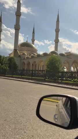 The video shows a view from the driver's side mirror of a car as it travels along a paved road. The mirror reflects the side of the car and the road ahead. In the background, a large mosque with a golden dome and four tall minarets is visible. The mosque is surrounded by trees and a decorative fence. The sky is blue with scattered white clouds, and the sun is shining. The scene appears to be in a city or town.