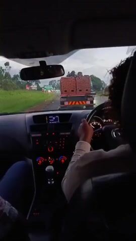 A first-person perspective video shows the interior of a Mazda vehicle with two occupants. A person with curly hair is behind the wheel, driving on a road. A sign indicating directions to Marondera and Grasslands is visible on the side of the road. Ahead, a large truck is carrying a load of bricks. The weather appears overcast. The vehicle's dashboard is illuminated with red and blue lights.