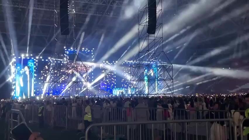The video shows a wide shot of a concert venue with a large crowd in the foreground separated by metal barriers. In the background, a massive stage is illuminated with numerous bright white and blue lights, creating beams that stretch across the venue. There are large screens on the stage displaying performers, though they are not clearly discernible. Several tall speaker towers are visible on either side of the stage. Smoke or haze is present in the air, catching the light. The overall atmosphere is that of a large-scale live music event.