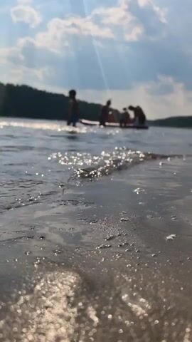 The video shows a group of people on a raft on a lake. The camera is close to the water, showing ripples and bubbles. The sun is shining through the clouds, creating light rays. One person on the raft is throwing water into the air.