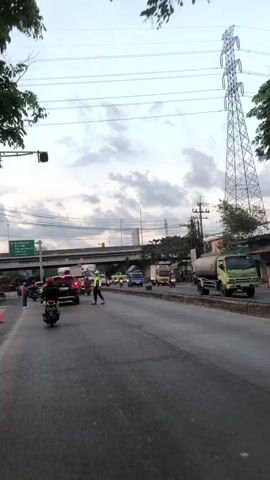 A traffic officer is directing traffic on a road. The officer is wearing a bright yellow vest and dark pants. The officer is standing in the middle of the road, with their arms extended. There are several vehicles on the road, including cars, motorcycles, and a truck. A pedestrian is crossing the road. The sky is cloudy. A large electrical tower is visible in the background. There is an overpass in the background.