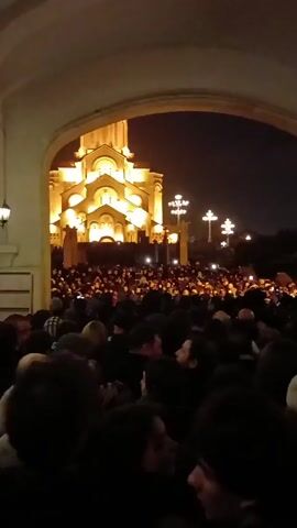 The video depicts a large crowd of people gathered outside a brightly illuminated church at night. The crowd is densely packed, and many individuals are looking towards the church. The church, a prominent building with multiple levels, is lit up in warm, golden tones. Streetlights are visible near the church, also contributing to the overall illumination. The shot is framed by an arched entryway, giving a sense of looking out from a covered area. The atmosphere appears to be active and potentially celebratory or religious.