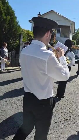 A marching band, dressed in black pants, white shirts, and black hats, walks down a cracked, sunlit street in a neighborhood. A person in the middle plays a brass instrument, seemingly following sheet music. Houses with small yards are visible on either side of the street, and parked cars are scattered along the curb.