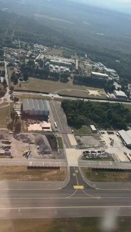This aerial video captures an airfield in Somma Lombardo, Italy, on a clear, sunny day, likely mid-morning or early afternoon given the bright sunlight and distinct shadows. The scene is dominated by an airport runway in the foreground, marked with yellow lines and numbers, including "65" and "MAX SPAN 36M".

Beyond the runway, the airfield grounds are visible, featuring various industrial buildings, hangars, and open areas with parked vehicles, construction materials, and what appear to be aircraft. A large, multi-bay hangar with a silver, corrugated roof stands out prominently. Adjacent to this, a smaller building and an area with a pile of aggregate are present. Further back, a cluster of red-tiled buildings suggests older infrastructure, possibly administrative or residential, surrounded by trees.

The mid-ground showcases a green forested area that borders the airfield, transitioning into a more developed area with larger buildings, possibly a business park or industrial complex, situated further in the distance. A road network is visible connecting these areas, with cars and trucks indicating ongoing activity. The background is filled with rolling, densely wooded hills, characteristic of the Italian landscape, under a bright blue sky.

There are no people explicitly visible in the video, but the presence of vehicles, construction materials, and the overall operational appearance of the airfield imply human activity. The video seems to be taken from an aircraft in the process of landing or taking off, as indicated by the slight angle and the reflection of what might be a window in the bottom right corner of the frame. The overall mood is one of industrial activity within a natural, picturesque setting.