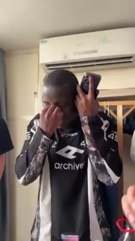 A person with dark skin is holding a smartphone up to their ear, as if on a call, while rubbing their eye and scrunching up their face. They are wearing a black and gray long-sleeved shirt with a white logo that says "archive" on it, as well as the number "45". In the background, another person is visible, wearing a black shirt with a pink heart graphic. The setting appears to be indoors, possibly a room with an air conditioning unit mounted on the wall, and a wooden coat rack with clothes hanging on it in the background.