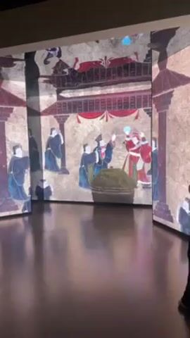 The video shows an interactive art exhibit. The artwork is projected onto three large rectangular panels, creating an immersive experience. The artwork depicts a scene from ancient China, featuring figures in traditional clothing and a structure resembling a temple or palace. Some figures appear to be gesturing or interacting with each other, while others stand to the side. The overall color palette is muted, with touches of red and blue highlighting certain elements. The panels are arranged to form a corner of a room, and the floor is dark and reflective. One person stands near the edge of the right-hand panel.