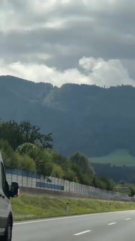 The video is shot from the perspective of a vehicle traveling on a highway, with a white van visible in the foreground on the left. The van is stationary at the beginning of the clip and then begins to move as the camera captures the highway. The highway stretches ahead, with other vehicles present, including cars and trucks. To the right of the highway, a sound barrier covered in greenery lines the road. Beyond the sound barrier, a verdant hillside rises, covered in trees and dotted with patches of grass. In the background, a mountain range with a dark, forested slope is visible, partially obscured by clouds. The sky is overcast with dramatic clouds, and hints of sunlight break through in some areas. The overall scene depicts a highway journey through a scenic, mountainous landscape.