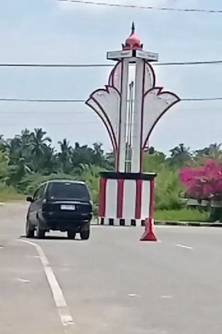 A black SUV drives past a monument on a sunny day. The monument is tall and has a red dome on top. It is white with red and white striped accents at the base. A traffic cone is placed on the road near the monument. The background consists of green trees and a blue sky.