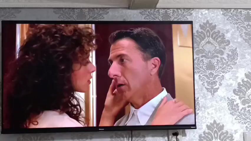 The video shows a close-up of a television screen displaying a scene from a movie. On the left of the screen is a woman with dark, curly hair and on the right is a man with short, dark hair, wearing a light-colored suit. The man appears to be speaking, with his mouth open, and the woman is looking at him, with her hand on his chest. The background of the scene appears to be indoors, with a patterned wallpaper visible on the wall behind the television. The lighting in the scene is warm and intimate. The image is a still frame, so no actions are occurring within the scene itself.