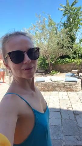 This video appears to be a selfie taken by a woman wearing a blue tank top and sunglasses. She is outdoors, possibly on a terrace or walkway paved with flagstones. In the background, there is lush green foliage, a stone wall, and a clear blue sky. To the left, another woman in a yellow top and shorts is visible, holding a red bag and taking a photo with her phone. The scene suggests a sunny, warm day in a scenic location, possibly a European coastal town.