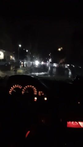 The video shows a view from inside a car at night, looking through the windshield. It is raining, and the windshield is wet with water droplets. The headlights of other cars and streetlights are visible through the rain, creating blurry, bright streaks on the wet glass. The car's dashboard is visible on the right, with the speedometer and various indicator lights illuminated. The overall scene is dark and suggests a rainy night drive in an urban or suburban area.