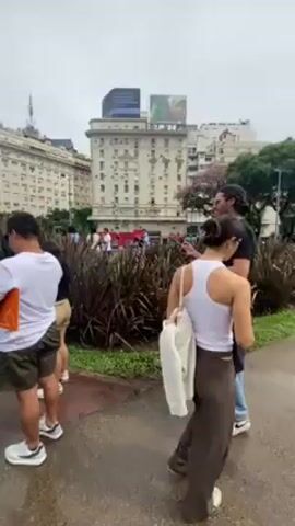 The video shows a group of people on a city street, standing on a red brick path. In the background is the Obelisco de Buenos Aires, a tall, white obelisk. Next to it is a large, green bush shaped into the letters "BA." The people are dressed in casual clothing and appear to be tourists or visitors taking pictures of the landmark. There are buildings in the background as well. The sky is overcast, suggesting it might be a cloudy or rainy day. The overall impression is that this is a tourist spot in Buenos Aires, Argentina.