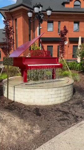 The video shows a bright red grand piano sculpture that functions as a fountain. Water cascades down from the piano's keys and the area where the lid would open, falling into a circular, stone-lined basin. The sculpture is situated outdoors in what appears to be a garden or landscaped area. Behind the fountain, a modern building with orange brick siding and dark roofing is visible. Several small, slender trees with dark red foliage stand on either side of the piano sculpture. A lamppost with multiple lights is positioned behind the sculpture. The ground around the fountain is covered with dark mulch and some loose soil. The overall scene suggests an artistic and decorative outdoor installation. There are no people clearly visible in the frame.