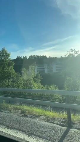 The media file captures a daytime drive through a scenic landscape, likely in Kongsberg, Norway, given the context. The scene is dominated by lush green trees and a clear blue sky with wispy clouds, suggesting fair weather. A road guardrail runs along the foreground, indicating the perspective is from a vehicle moving along a road or highway.

In the background, a multi-story building with a modern architectural style is visible amongst the trees, suggesting a developed area within a natural setting. The sun is shining brightly, casting lens flare and illuminating the scene, which implies it's likely late morning or early afternoon.

One notable detail is a set of road signs. The first sign, appearing around 00:02, is a triangular "Give Way" sign with a red border, commonly seen at intersections. Below it is a circular sign indicating a roundabout 200 meters ahead, also confirming traffic flow information. Another signpost is visible at 00:03.

The overall impression is one of a peaceful journey through a visually appealing environment, with the road signs providing practical navigational context. There are no people visible in the media. The activity is simply traveling by vehicle.