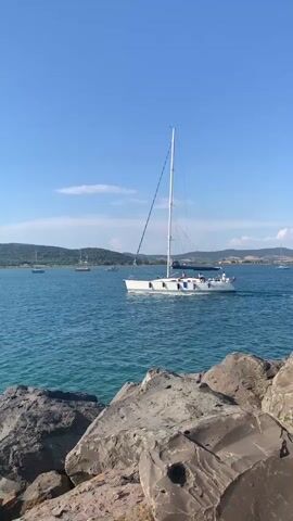 A white sailboat with a single mast sails across a body of blue water. The boat has several people visible on deck. In the foreground, large gray rocks form a barrier. In the background, green, tree-covered hills meet a clear blue sky. The sky is largely cloudless, with a few wispy clouds scattered near the horizon. The water is calm, with gentle ripples on the surface. The lighting suggests it is daytime, with bright sunlight.