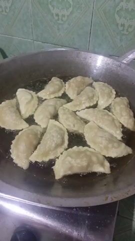 The video shows a close-up shot of a wok filled with hot oil where several crescent-shaped pastries are being deep-fried. The pastries are arranged in a single layer, with some overlapping. They appear to be light in color, suggesting they are either uncooked or very lightly fried. The oil is bubbling around the pastries, indicating it is hot. The background is blurred but shows a tiled wall in green and white. The camera is positioned slightly above the wok, looking down at the frying process.