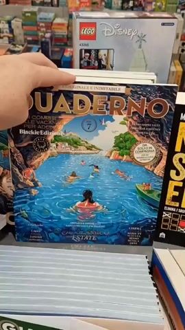 The video shows a person's hand holding a magazine with a colorful cover depicting a scenic bay with people swimming and a boat. The magazine is open, revealing some of its pages. In the background, shelves are filled with books and toys, including a Lego Disney set. Another book, titled "NON È STATO EMM..." is partially visible to the right of the magazine. The hand is turning the pages of the magazine, showing illustrations and text. The overall environment appears to be a bookstore or a retail space.