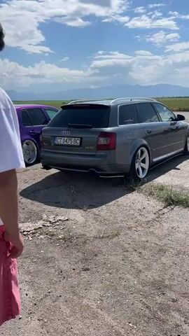 The video shows a man in a white t-shirt and red pants standing in front of two cars parked in a dirt lot on a sunny day. One car is a purple Volkswagen Golf and the other is a gray Audi A4 Avant. The Audi is idling and emitting a large amount of black exhaust from its tailpipe. Several other cars are parked in the background. The landscape is a flat field with mountains in the distance under a partly cloudy sky. The man walks towards the camera, partially obscuring the cars, then moves slightly to the left to provide a view of the cars and smoke again.