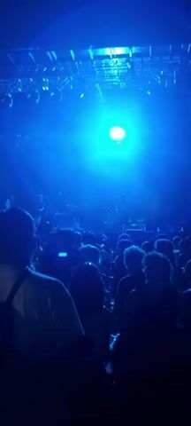 The video shows a dimly lit concert venue. The stage is visible in the background with a band performing. Lights, primarily blue, illuminate the stage and the audience. The audience is silhouetted against the lights, with some individuals holding up phones to record. The video is shaky, suggesting it was recorded by someone in the audience.
