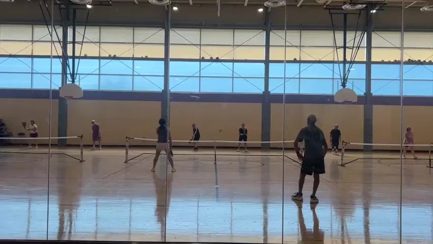 A group of people are playing pickleball indoors. The court has a wooden floor and is surrounded by glass walls. A net divides the court in half. Several players are actively engaged in hitting a ball with paddles, while others are observing or waiting for their turn. The room appears to be a gymnasium with high ceilings and windows.