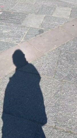 The video begins with a shadow cast on a paved surface, then transitions to a wide shot of a large, ornate cathedral. The building has a red-brown stone facade with numerous arched windows, spires, and decorative elements. Several people are visible in the plaza in front of the cathedral, some standing and walking, others sitting on benches. The sky is clear and blue. The camera pans slightly, showing different angles of the building.