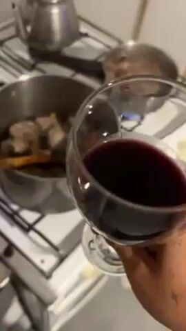 A hand holds a wine glass filled with red wine in front of a stove. On the stove, a pot with meat and vegetables is being stirred with a wooden spoon, while a pan on another burner shows some bubbling liquid. A silver kettle is also on the stove. The hand holding the wine glass has a tattoo on it.