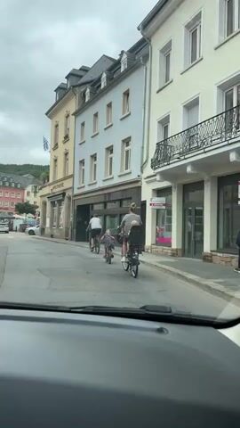 The video shows a family on bicycles riding down a street in a European town. A person is driving a car, and the video is filmed from inside the car, looking forward. The street is lined with buildings. On the right side of the street, there is a building with a sign that says "APOTHEKE." Further down the street, there is a town square with more buildings and outdoor seating for cafes. The sky is overcast.