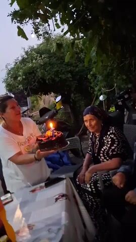 A woman in a white shirt holds a chocolate birthday cake with lit candles towards an elderly woman wearing a headscarf and a patterned dress. The elderly woman is seated at a table. To her right, a man in a checkered shirt is also seated. Behind the woman with the cake, several men are visible; one is holding up a phone, seemingly recording. Another man in a yellow tank top is also present. The setting appears to be outdoors, possibly a backyard, with trees and foliage in the background. The lighting suggests it is dusk.