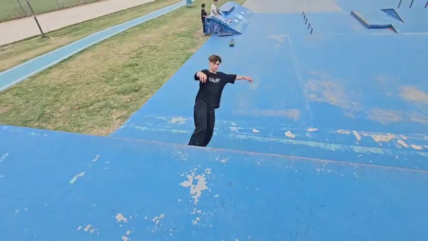 A young man on a skateboard, wearing a black shirt and pants, skates down a blue ramp at a skateboard park. Other people are scattered around the park, near ramps, railings, and benches.  The area appears to be outdoors with a patch of green grass and trees visible. The skateboard park is painted with blue and has white markings. Another young man, wearing a black shirt and shorts, is skating nearby. He has a hat on backwards.