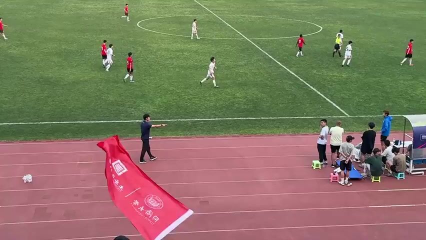 A soccer game is in progress on a green field, viewed from an elevated position overlooking the track surrounding the field. Players in red and white uniforms are scattered across the field.  A person standing on the track is waving a red flag with white writing on it, seemingly supporting one of the teams. The flag features a university logo and the words "COLLEGE OF CIVIL ENGINEERING". Behind the flag waver, several people are sitting on small stools, likely part of a team support group. In the background, there are buildings, suggesting an urban setting. The scene conveys the atmosphere of a lively sporting event with team spirit.