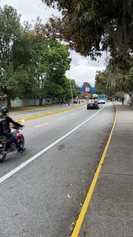 The video shows a view from a sidewalk of a multi-lane road with traffic. A motorcycle with a black delivery box on the back is in the foreground, moving away from the camera. Behind it, several other motorcycles and cars are visible, as well as a bus further down the road. The road is bordered by yellow lines on the right and has a white line separating the lanes. On the left side of the road, there are trees and some buildings with colorful graffiti. The trees on the right side of the road have hanging moss. The sky is overcast. There are traffic signs visible in the distance.