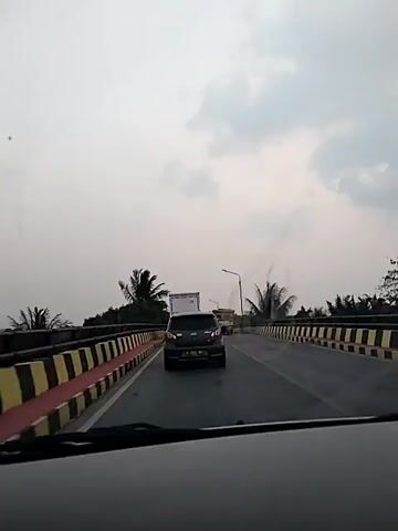The video shows a view from a car driving on a road. The road has yellow and black striped barriers on the sides. There are several vehicles on the road, including cars and trucks. Palm trees are visible on the left side of the road. The sky is overcast. The video appears to be shot during the day.