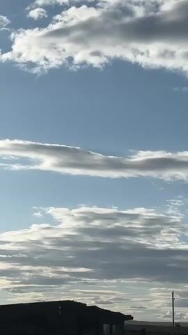 The video shows a view of the sky with clouds and the sun. The scene starts with a view of light blue sky with small, scattered white clouds. Then the camera angle shifts upwards revealing many clouds. The clouds appear layered and are gray and white in color. The sun is visible behind the clouds, emitting a bright light. In the foreground, there is a silhouette of a building. There is also a telephone pole with wires. The video is filmed from a low angle, emphasizing the sky and clouds.