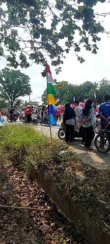 A group of people are gathered on the side of a road. Many of them are on motorcycles. There is a tall pole with a colorful flag that appears to be made of multiple rectangular pieces of fabric in red, white, green, and yellow. Several people are standing near the pole. The road appears to be paved. There are trees with green leaves overhead. In the foreground, there is a ditch with dry dirt and grass. Some people are wearing headscarves. Several motorcycles are parked and in motion. The overall impression is of a gathering or a procession.