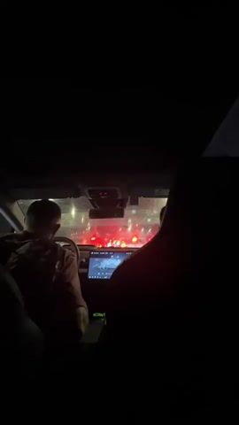 The video shows the interior of a car at night. A person is visible in the driver's seat, facing forward. The dashboard display is illuminated. Through the windshield, there are red lights that appear to be from traffic signals or other vehicles. The overall lighting is dark, with reflections visible on the windshield.