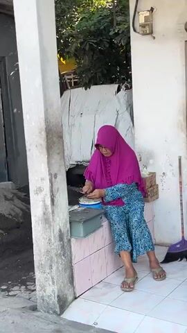 A woman wearing a purple hijab and a blue and green patterned dress sits on a pink tiled ledge in front of a building. She is using a knife to cut something on a white plate. To her left, there is a grey bin and a pile of dark material. To her right, a broom leans against the wall, and a black cat is partially visible near the broom. The background shows a white wall, foliage, and parts of a building. The lighting suggests it is daytime with sunlight.