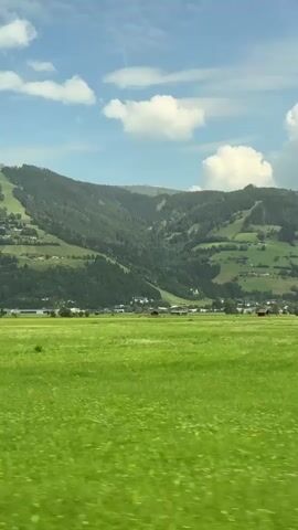The video captures a panoramic view of a vibrant green meadow under a bright blue sky with scattered white clouds. In the background, lush green mountains rise, dotted with trees and small houses, suggesting a rural or mountainous landscape. The sun appears to be shining, casting a natural light on the scenery. The perspective suggests the video was recorded from a moving vehicle, possibly a train or car, as indicated by the slight blur and the continuous panning motion across the landscape. The overall atmosphere is serene and picturesque, showcasing a natural environment with signs of human habitation.