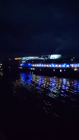 A nighttime video shows a large boat with blue lights along its side, moving across dark water. The lights from the boat reflect on the water's surface, creating a shimmering effect. In the background, other distant lights are visible, suggesting a waterfront area or cityscape. The sky is dark with some clouds. There are no people visible in the video. The boat appears to be in motion, indicated by the wake it is creating.