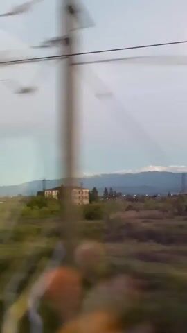 A slow-motion shot from a moving vehicle captures a distant landscape under a pale sky. The view, seen through a slightly smudged window, shows a cluster of buildings nestled among lush green trees. In the background, majestic mountains rise against the horizon, their peaks softened by distance. The scene is quiet, with no people or animals visible, only the muted beauty of the natural world and human structures blending together. The overall atmosphere is one of peaceful observation, a fleeting glimpse of a tranquil rural setting.