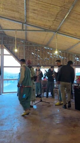 A group of men is gathered inside a building with a thatched roof, apparently performing live music. They are dressed in traditional Moroccan clothing. Several men are holding stringed instruments, and there is a microphone in front of them, suggesting they are singing or speaking as well. One man, dressed in green and yellow, is dancing. A sign on a small table reads "TIPS".  A large window in the background offers a view of a beach or coastline. The setting appears to be casual, possibly a restaurant or gathering place.  Several people are seated and watching the performance.  The floor has a reddish-brown mottled look.