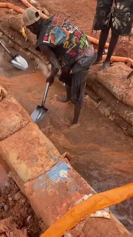 In a scene of manual labor in Rayfield, Nigeria, two individuals are engaged in what appears to be construction or excavation work. The ground is a muddy, reddish-brown earth, suggesting recent rainfall or an active construction site.

The primary subject, a person wearing a black t-shirt with a colorful graphic print, a dark skirt or wrap, and a baseball cap, is partially submerged in muddy water. They are holding a shovel and seem to be actively clearing debris or digging. Their hands are covered in mud, indicating strenuous activity.

Another individual, standing to the right, is partially visible, wearing shorts and appearing to observe or assist. Their bare feet are also in the muddy terrain.

Several large sandbags are strategically placed, forming a barrier or a pathway, indicating an effort to manage water or soil. Orange pipes are also visible, suggesting the presence of water management or supply systems. The overall impression is one of hard work in challenging conditions, likely to be during daylight hours given the brightness of the scene. The activity appears to be practical and physically demanding.