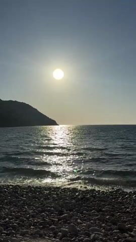 The video shows a daytime view of a rocky beach and the ocean. The sun is bright in the sky, casting a reflection on the water. There is a small hill visible in the background. The tide is gentle, with small waves lapping at the shore. The sky is clear and blue.