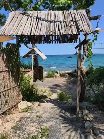 The video pans across a rustic entrance to a beach. The entrance is framed by a thatched roof supported by wooden posts, with bamboo fencing on the left. Beyond the entrance, a path leads to a rocky shoreline with clear turquoise water. Several small boats are visible on the water, and the coastline with buildings is visible in the distance under a bright blue sky. String lights are hung across the scene, adding a touch of ambiance. The overall impression is one of a simple, natural, and inviting beachside location.