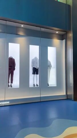 The video shows a retail store window display featuring mannequins dressed in casual apparel. The window is divided into illuminated vertical sections, each showcasing a different outfit. The brand name "BOSIDENG" is visible at the bottom of the display. The floor in front of the store is blue with a wavy pattern. The video pans to the right, revealing more of the store's exterior, including a logo and seating area with chairs and a table. Text appears on the glass, including "AREAL KIM JONES BOSIDENG" and Chinese characters.