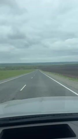 The video shows a vehicle driving on a gray asphalt road. The sky is overcast. On the left side of the road is a green field, and on the right side is a brown, plowed field. A row of trees runs along the horizon. The camera is positioned inside the vehicle, showing the dashboard. The dashboard display indicates a speed of 103 km/h. The car appears to be in autopilot mode. The video ends with a view of the steering wheel.